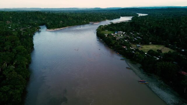 A drone aerial view looks across a broad river bordered by dense tropical rainforest near Misahualli, Ecuador. Ripples texture the blue water beneath a bright, cloud-streaked sky, while dark tree-line