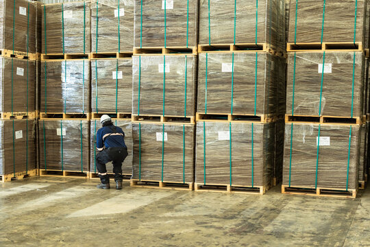 A Black inspector does inventory along a wall of shrink wrapped pallets in a large storage hall, documenting skilled work in the era of AI and robotic automation