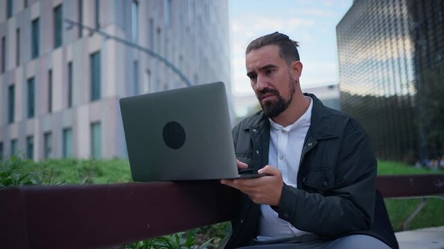 A business man seated by an outdoor bench studies his laptop screen, pauses with a hand on his chin, then resumes using the computer.
