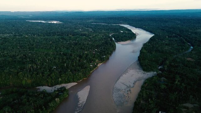 A broad aerial panorama shows a muddy river curving through dense tropical forest and past the riverside town of Misahualli, Ecuador. Green hills and distant ridgelines sit beneath low clouds, with ro