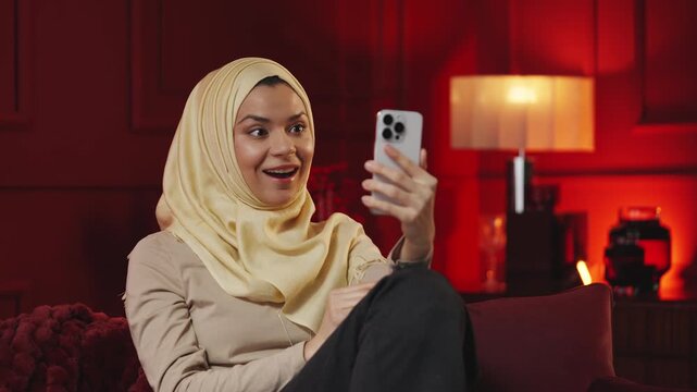 Cheerful Muslim woman in hijab having video call on phone while at home