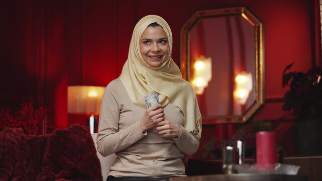 Young happy Arab woman in hijab counting cash, saving for dream purchase
