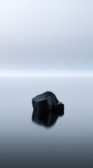 Fototapeta premium A solitary dark rock reflecting peacefully on the smooth, misty water surface under a soft gradient sky in a minimalist and tranquil natural setting