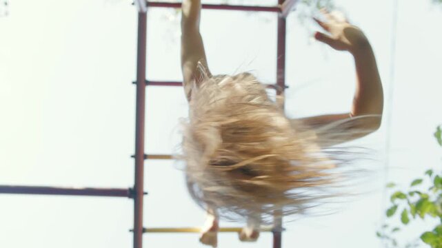 Joyful child swings upside down on monkey bars at sunny park, enjoying playful outdoor activity. Smiling young girl shows carefree emotion while hanging above camera in summer sunlight.