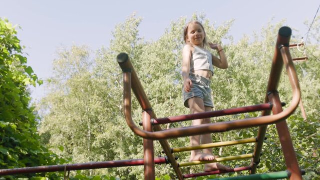 Cheerful school age girl navigates rusty playground climbing frame during bright summer day. Active kid carefully steps down steel ladder rungs, enjoying energetic outdoor recreation in park.