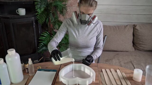 Woman wearing respirator mask and gloves pours white resin into heart-shaped silicone mold on wooden table during tabletop casting in indoor craft workshop