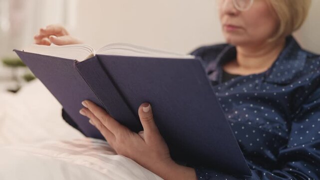 Focused woman in her 50s reads historical book while lying in bed at home