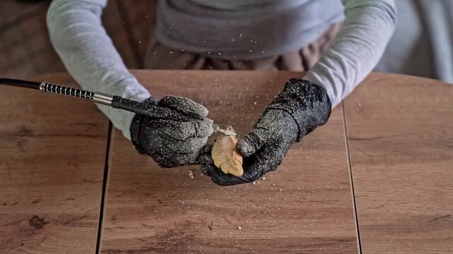 Close overhead view of gloved hands carving wooden leaf with rotary tool, shaping detailed craft piece as fine wood dust and shavings scatter on table