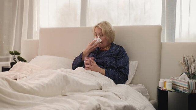 Sad middle-aged woman coughs in bed while holding hot drink, suffering from flu