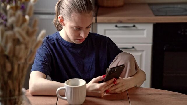 Young woman sitting at kitchen table scrolling smartphone with coffee mug on wooden tabletop, natural window light and casual home interior, focused candid lifestyle scene