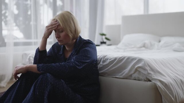 Unhappy middle-aged woman in pajamas sits on floor, struggling with depression