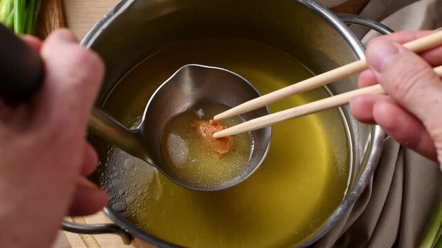 Chopsticks dissolve shiro miso in broth inside a ladle held above a pot in a close overhead view.