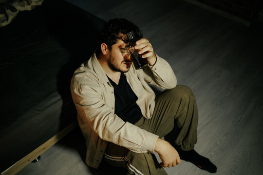 Exhausted youth rests alcohol bottle against face in dark bedroom. Surrendering to deep grief
