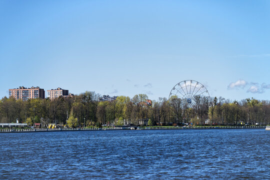 Panoramic waterfront of upper pond in Kaliningrad with calm water, tree-lined shore, autumn foliage and distant Ferris wheel under bright clear blue daytime sky, Russia
