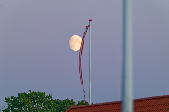 Evening sky with moon rising behind a flagpole and rooftop with trees
