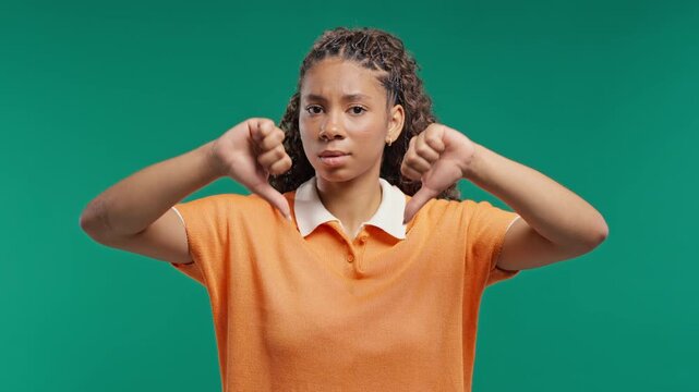 Teenage lady expressing discontent with showing thumbs-down gesture on green studio. Unhappy african american woman condemns with dislike.