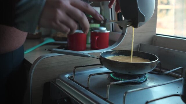 Female hands close up pouring beaten eggs from a bowl into a small frying pan on a gas stove in a campervan kitchen.