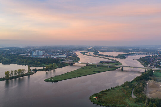 Aerial view of the Claus power plant along the Meuse river, featuring industrial facilities, a bridge, and greenery under a soft sunset sky in Maasbracht, Limburg, Netherlands.