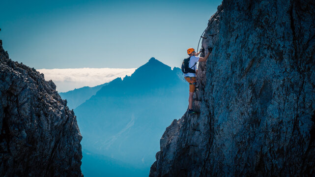 Solo male climber ascending a vertical rock wall on a via ferrata in the Austrian Alps near Innsbruck, Tyrol, with rugged cliffs and distant mountain peaks creating a dramatic alpine scene.