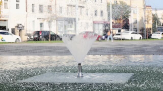 Water fountain spraying in city square on sunny day with buildings and cars in blurred background