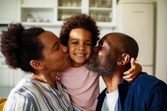 Parents kiss smiling child on both cheeks at home