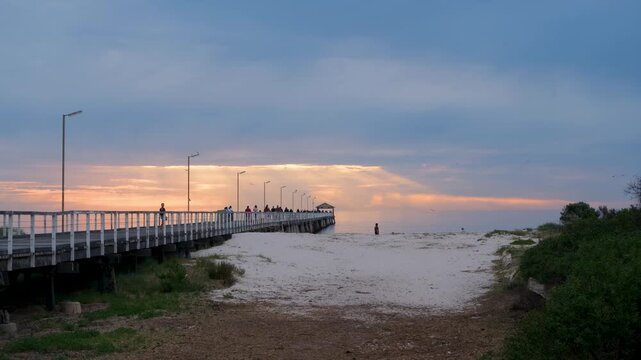 Silhouetted people walking along long Semaphore Jetty at sunset in Semaphore, Adelaide, South Australia, seagulls flying overhead. Seaside leisure and evening relaxation, a local tourist attraction