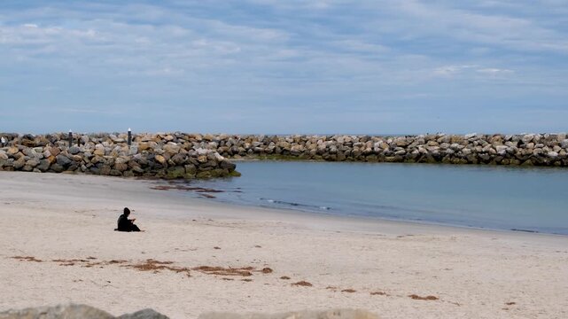 A person sitting alone on a sandy beach beside a rock breakwater at West Beach, Adelaide, South Australia, with calm sheltered water. Solitude and quiet reflection by the sea, personal moment.