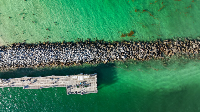 Aerial view of the Government Cut stone jetty and wooden pier surrounded by turquoise water with people swimming and walking on the pier in Miami-Dade County, Florida, United States.