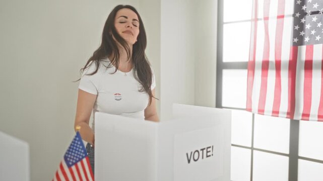Young brunette woman points finger to i voted sticker on her chest at a polling booth in a building with an american flag visible; civic pride.