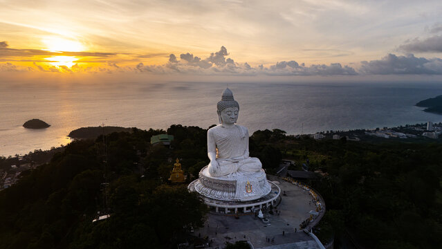 Aerial view of the Phuket Big Buddha statue on a hilltop overlooking the Andaman Sea at sunset with golden light reflecting on the water in Karon, Phuket, Thailand.