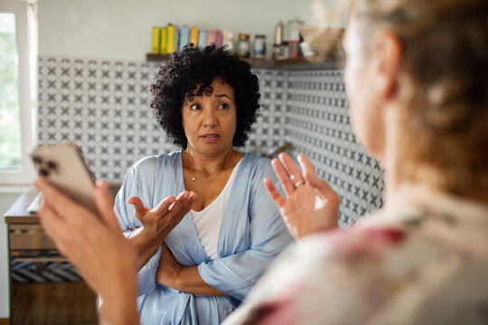 Woman shrugs at another woman showing phone in kitchen