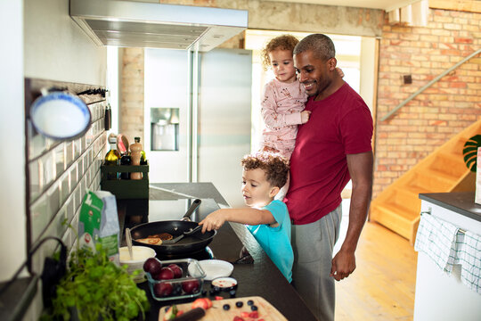 Father holds toddler while child flips pancake in home kitchen