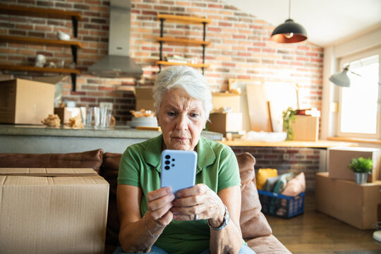 Senior woman looking at smartphone among moving boxes at home
