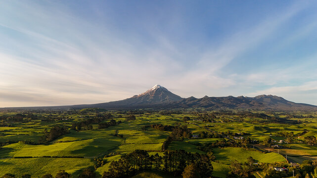 Aerial view of Te Papa-Kura-o-Taranaki with its snow-capped peak rising above the lush green rolling hills and farmland of the Taranaki Region, New Zealand.