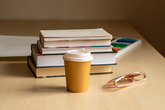 Takeaway coffee cup beside stacked books and eyeglasses on a desk. Studying and reading break. Education, learning and schoolwork. Student workspace with art supplies.