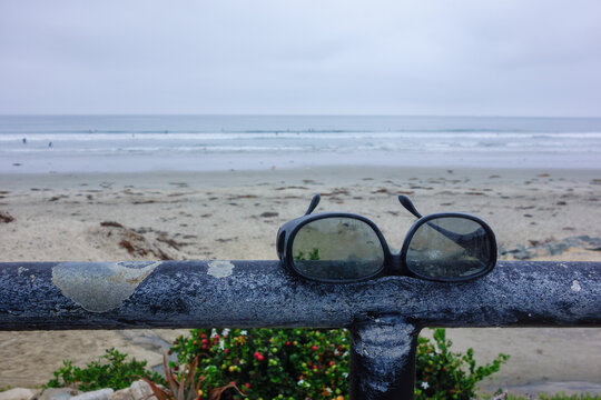 Black sunglasses rest on a metal railing overlooking a wide sandy beach with breaking ocean waves under overcast skies, Tourmaline Surf Park, San Diego, CA