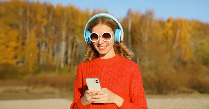 Happy young woman with phone enjoying listening to music in headphones outdoors in autumn park
