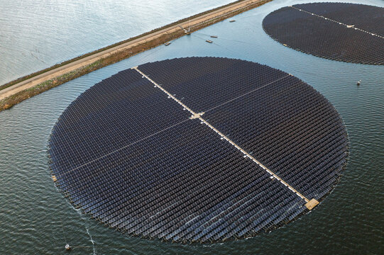 Aerial view of a circular floating solar park on the water, featuring rows of photovoltaic panels arranged in a large circle next to a coastal embankment in The Netherlands.
