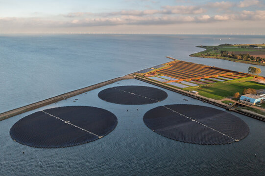 Aerial view of a floating solar park featuring three large circular solar arrays on the water, situated next to a coastal dike and green fields in The Netherlands.