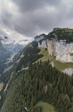 Aerial view of Aescher Guesthouse nestled into a sheer cliff face above dense pine forests in the Swiss Alps, with dramatic mountain peaks in Schwende-Rute, Appenzell Innerrhoden, Switzerland.