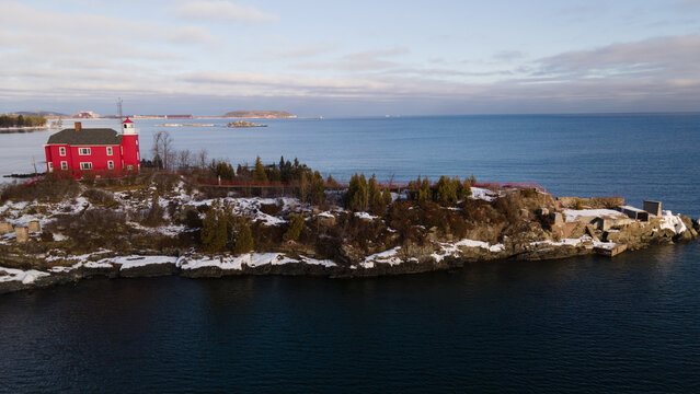 Aerial view of the red Marquette Harbor Lighthouse situated on a rocky, snow-dusted peninsula extending into the blue waters of Lake Superior in Marquette, Michigan, United States.