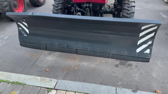 Tractor snow plow attachment with grey steel blade and striped safety markings stands on asphalt outdoors. Industrial vehicle equipment, heavy machinery part, commercial winter road service.