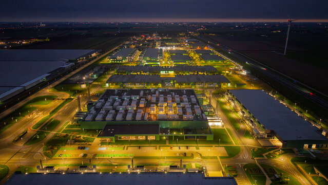 Aerial view of a large illuminated data center complex at night, featuring rows of industrial buildings, parking areas, and surrounding infrastructure in The Netherlands.