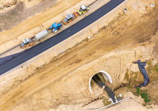 Aerial view of a road construction site with heavy machinery including rollers and a tractor on a new asphalt road next to a concrete culvert in Kartuzy, Pomeranian Voivodeship, Poland.