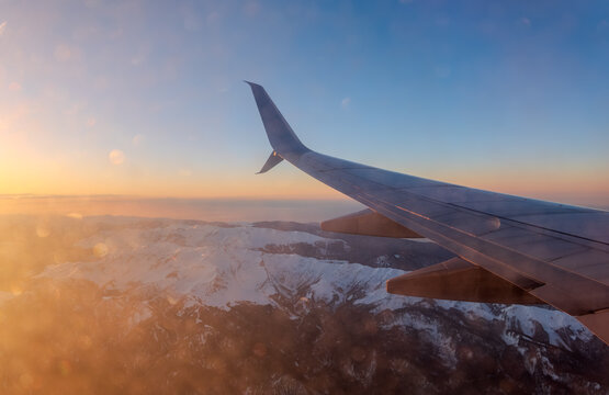 Stunning aerial view of mountain range at sunrise or sunset from airplane window, highlighting dramatic sky, clouds, and golden light on snow-capped peaks.