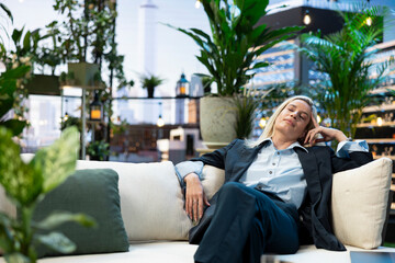 Fototapeta premium Businesswoman rests with eyes closed and one hand at her temple on a rooftop sofa after finishing work in a lush outdoor terrace setting.