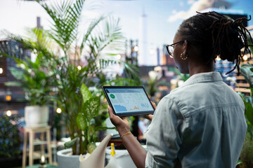 Woman monitors smart gardening app dashboard on tablet among potted plants in rooftop garden. Plant owner tracks vegetation live moisture readings to identify dry soil in need of watering. © DC Studio