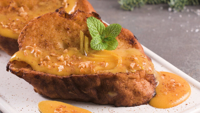 Traditional Christmas Rabanadas or Spanish Torrijas topped with creamy custard sauce, chopped nuts, and fresh mint on a white rectangular plate.