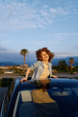 A cheerful woman leans on the roof of a car in a sunny outdoor setting, denim jacket over a striped top. A bright lifestyle moment captured for fashion and travel themes. © SHOTPRIME STUDIO