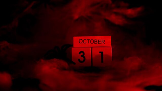 Dark still life composition featuring wooden block calendar displaying date 31 October Halloween holiday arrangement. Dramatic red lighting enhances mysterious atmosphere seasonal decorations props